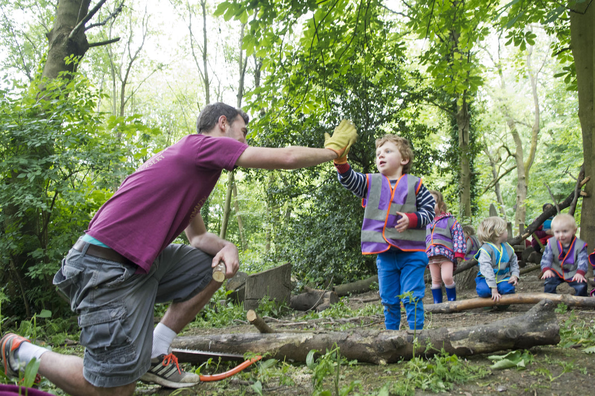 An adult with an arm raised beside a child looking up at them in woodland
