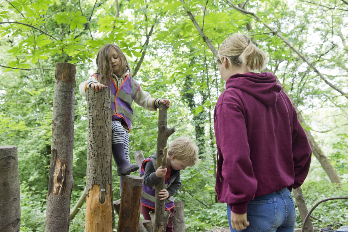 Children climbing on upright log stumps while an adult stands back and watches