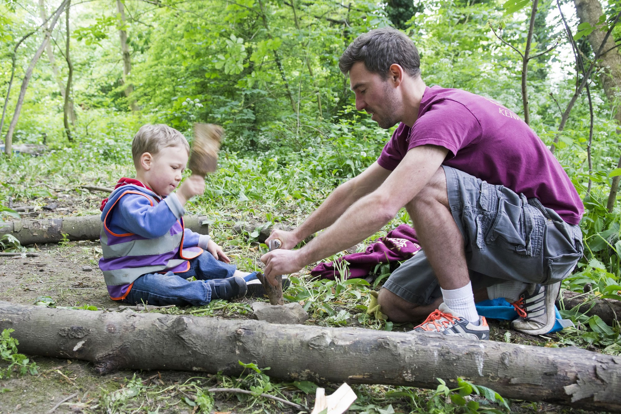 An adult kneeling beside two children working with hand tools on a fallen log
